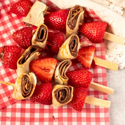 Close-up of strawberry and hazelnut crepe skewers arranged on red gingham parchment paper, featuring sliced rolled crepes with chocolate hazelnut spread and halved strawberries on wooden picks.