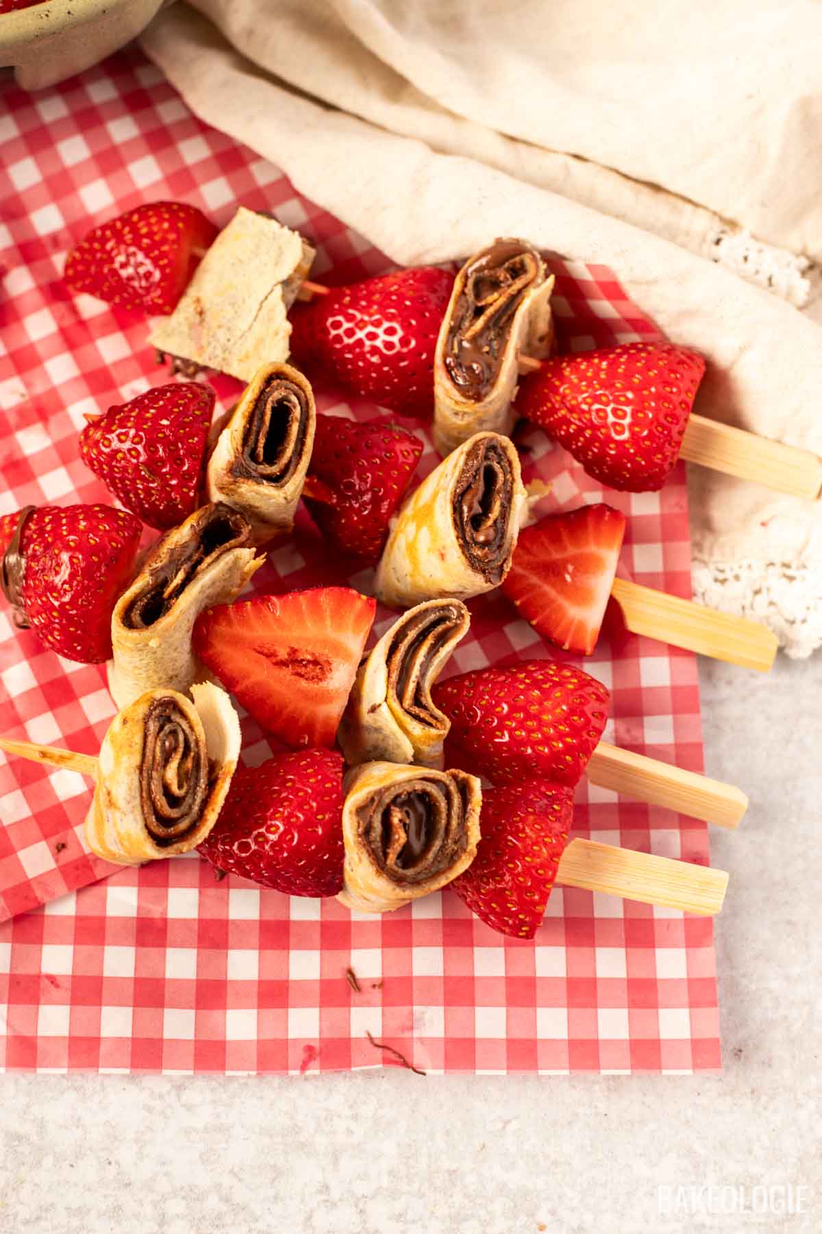 Close-up of strawberry and hazelnut crepe skewers arranged on red gingham parchment paper, featuring sliced rolled crepes with chocolate hazelnut spread and halved strawberries on wooden picks.