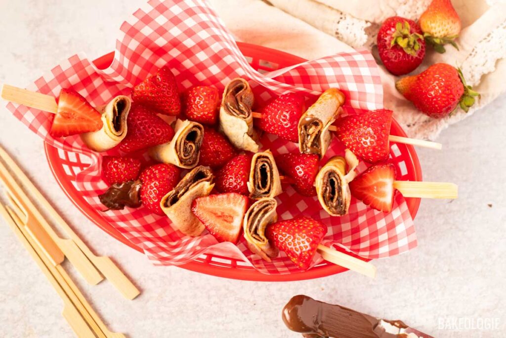 Top-down view of a red basket filled with strawberry and hazelnut crepe skewers, placed on red gingham paper with wooden skewers beside it and a swipe of hazelnut spread on the table, surrounded by loose strawberries and a linen cloth.