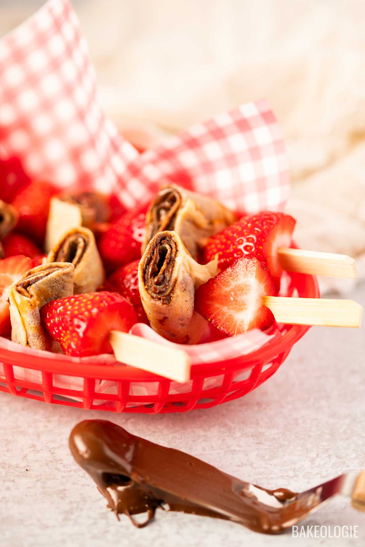 Close-up of strawberry and hazelnut crepe skewers in a red basket with red gingham liner, featuring rolled crepes and strawberries on wooden picks, with a swipe of chocolate hazelnut spread and a butter knife in the foreground.