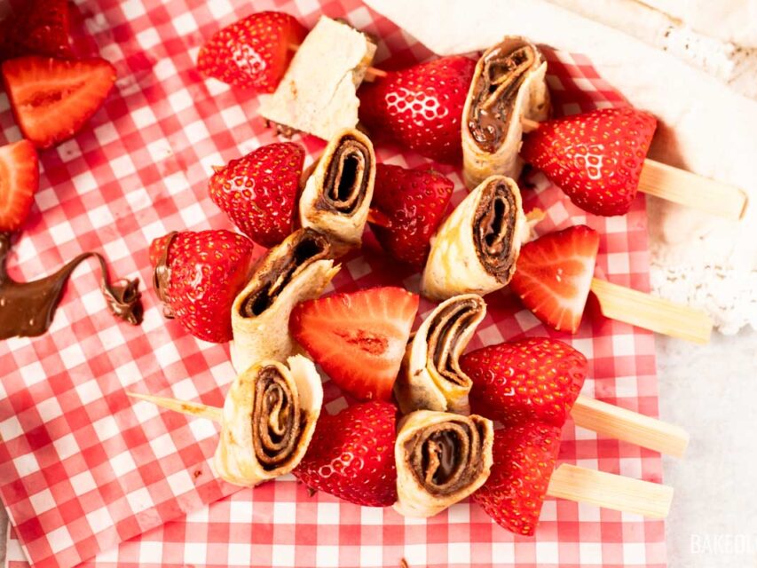 Close-up of strawberry and hazelnut crepe skewers on red gingham paper, showing rolled crepes with chocolate hazelnut spread alternated with fresh strawberries on wooden picks — a fun no-bake summer dessert.