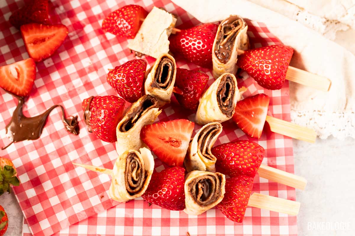 Close-up of strawberry and hazelnut crepe skewers on red gingham paper, showing rolled crepes with chocolate hazelnut spread alternated with fresh strawberries on wooden picks — a fun no-bake summer dessert.