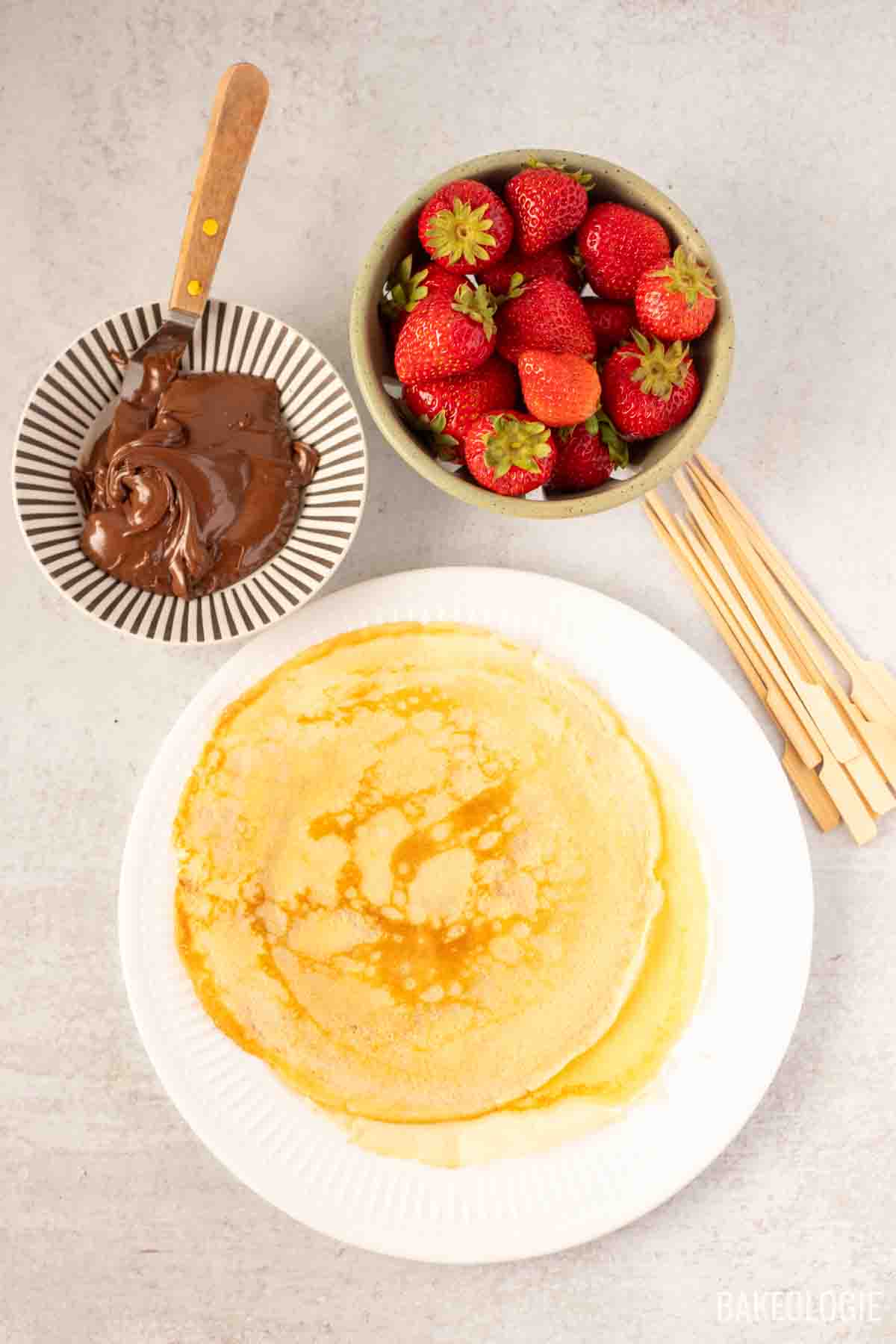 Flat lay of ingredients for strawberry hazelnut crepe skewers: a plate of stacked golden crepes, a bowl of fresh strawberries, a dish of chocolate hazelnut spread with a spreading knife, and a bundle of wooden skewers on a light countertop.