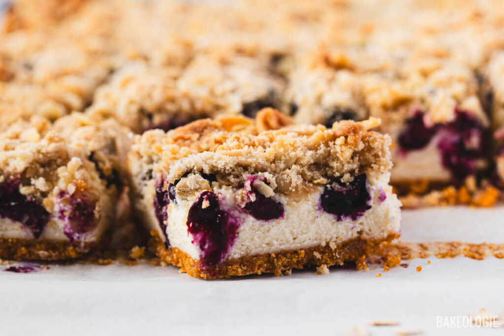 Sliced blueberry crumble cheesecake bars arranged on parchment paper, showing layers of crisp graham cracker crust, creamy cheesecake, juicy baked blueberries, and golden oat-almond crumble topping.