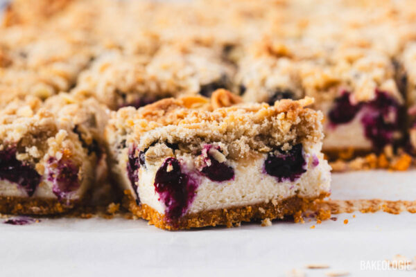 Sliced blueberry crumble cheesecake bars arranged on parchment paper, showing layers of crisp graham cracker crust, creamy cheesecake, juicy baked blueberries, and golden oat-almond crumble topping.
