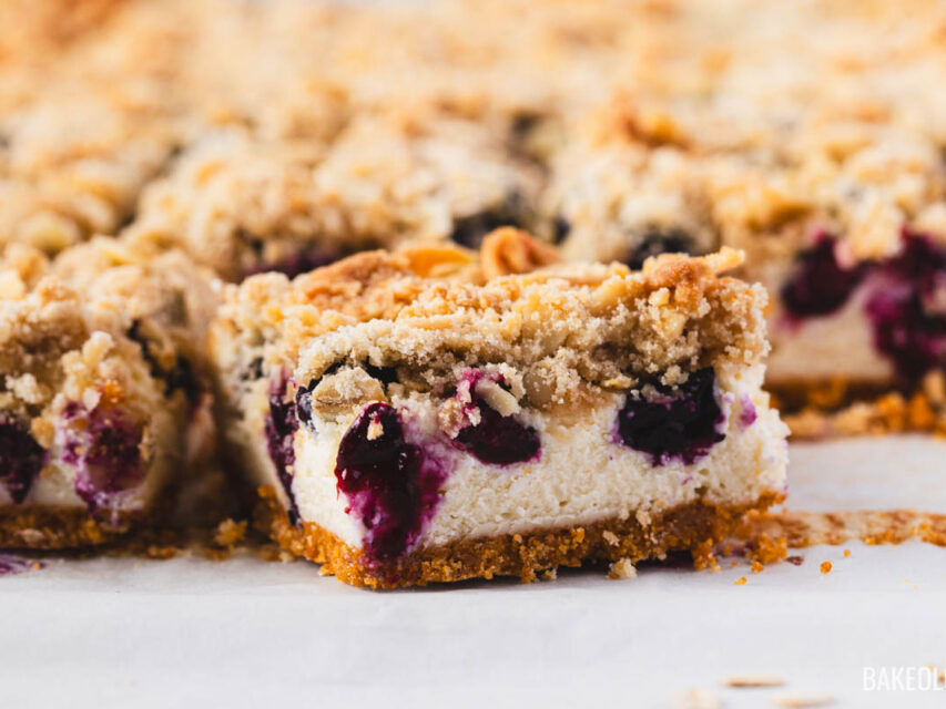 Sliced blueberry crumble cheesecake bars arranged on parchment paper, showing layers of crisp graham cracker crust, creamy cheesecake, juicy baked blueberries, and golden oat-almond crumble topping.