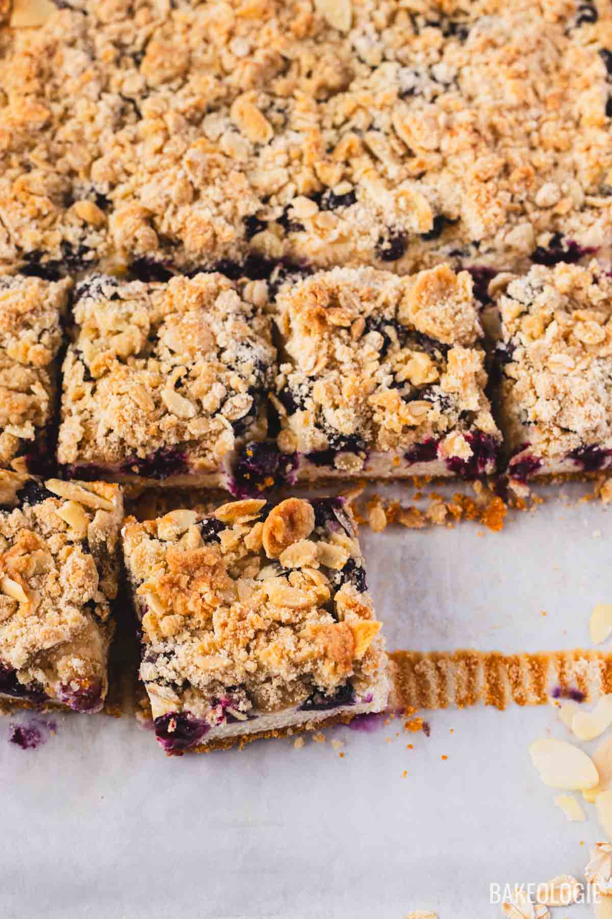Top-down view of freshly sliced Blueberry Crumble Cheesecake Bars on parchment paper, showing a golden almond oat crumble topping, juicy blueberry filling, and neat squares with buttery graham cracker crust.