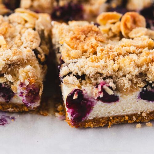 Close-up of a Blueberry Crumble Cheesecake Bar showing layers of buttery graham cracker crust, creamy cheesecake, juicy blueberries, and a golden almond oat crumble topping, with crumbs and blueberry filling on parchment paper.