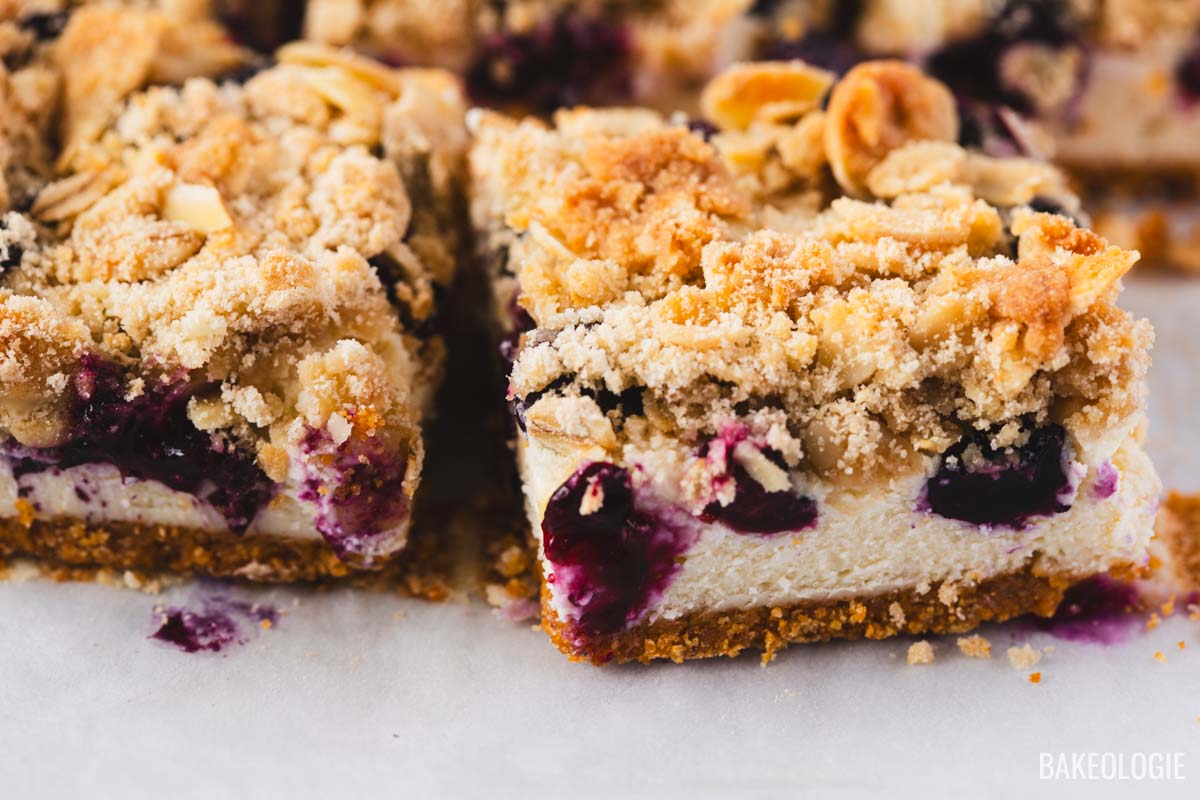 Close-up of a Blueberry Crumble Cheesecake Bar showing layers of buttery graham cracker crust, creamy cheesecake, juicy blueberries, and a golden almond oat crumble topping, with crumbs and blueberry filling on parchment paper.