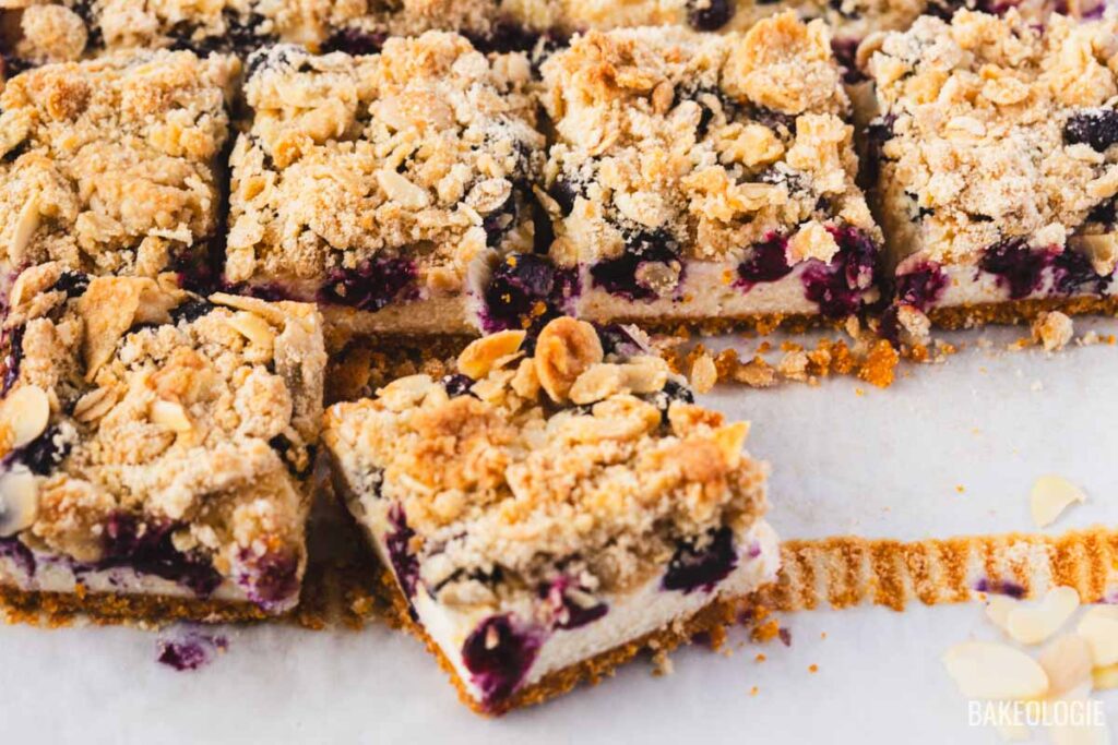 Sliced Blueberry Crumble Cheesecake Bars on parchment paper, showing neat layers of cheesecake, juicy blueberries, and golden crumble, with clean edges and crumbs scattered around.
