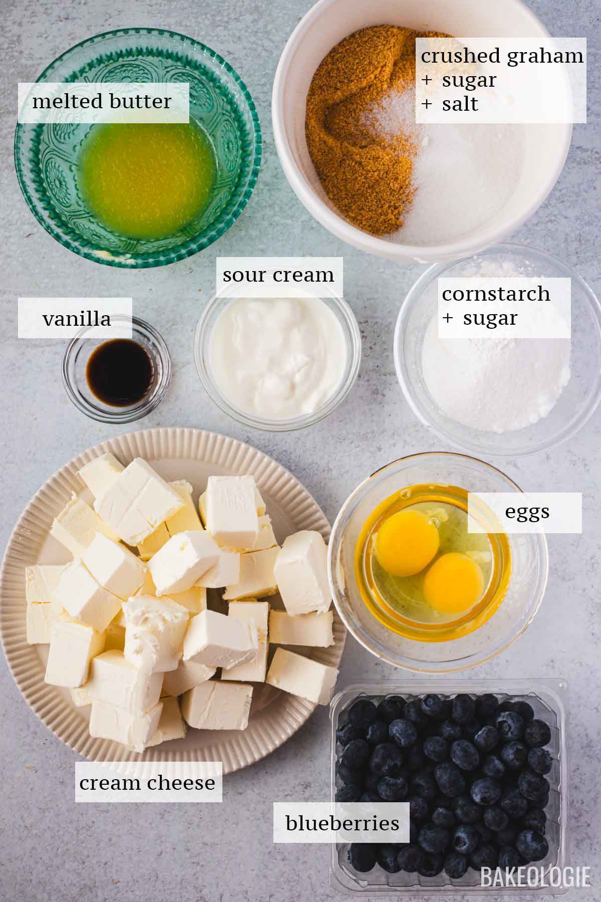 Overhead view of measured and labeled ingredients for blueberry crumble cheesecake bars, including cubed cream cheese, eggs, fresh blueberries, sour cream, melted butter, crushed graham crackers with sugar and salt, vanilla extract, and a bowl of cornstarch with sugar, all arranged on a light surface.