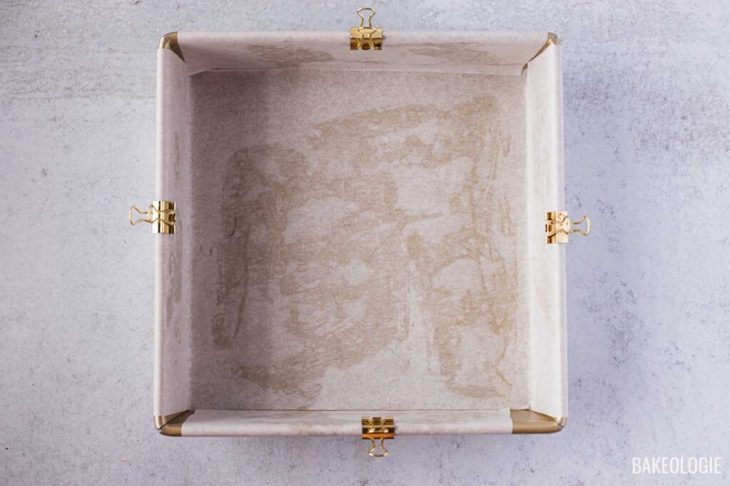 9-inch square baking pan lined with parchment paper and secured with gold binder clips, ready for making Blueberry Crumble Cheesecake Bars.