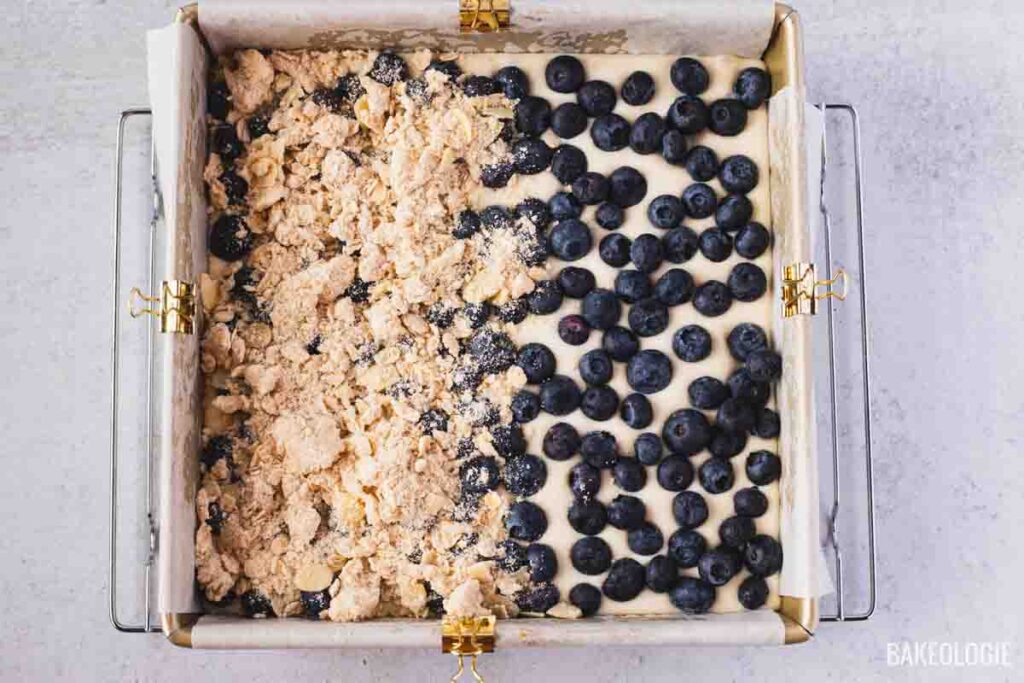 Top-down view of a parchment-lined baking pan half topped with fresh blueberries and half topped with almond crumble, showing the process of assembling Blueberry Crumble Cheesecake Bars before baking.