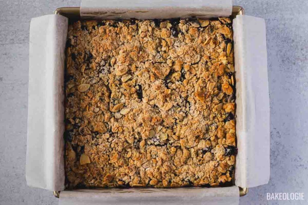 Overhead shot of freshly baked Blueberry Crumble Cheesecake Bars in a parchment-lined pan, showing a golden, crispy crumble topping with baked blueberries peeking through.