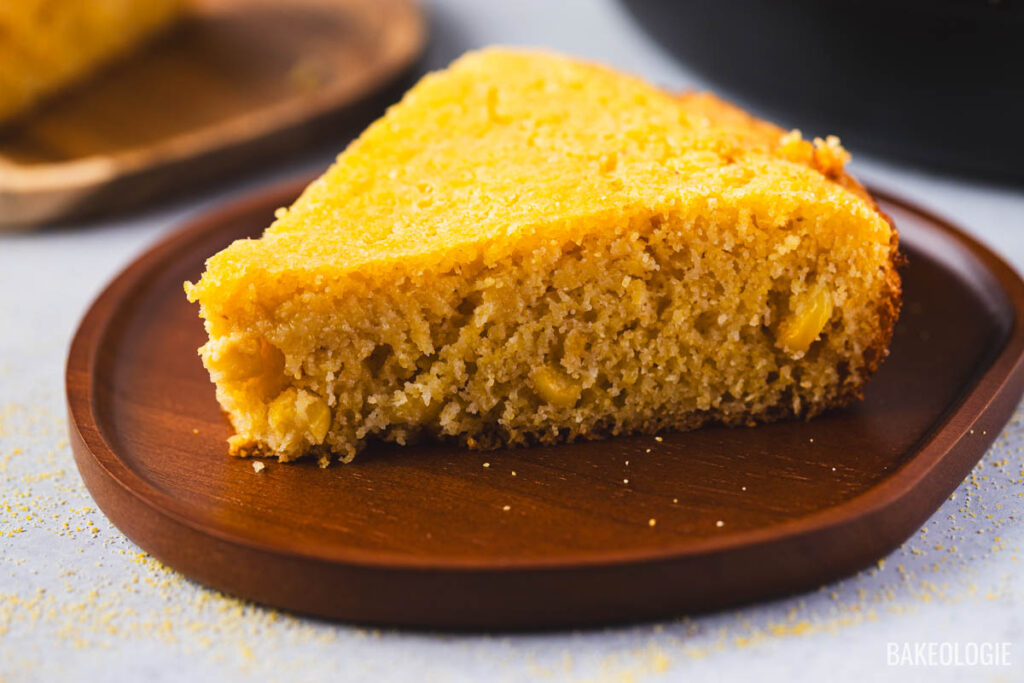 Slice of homemade skillet cornbread on a wooden plate, showing moist tender crumb with corn kernels and crispy golden edges.