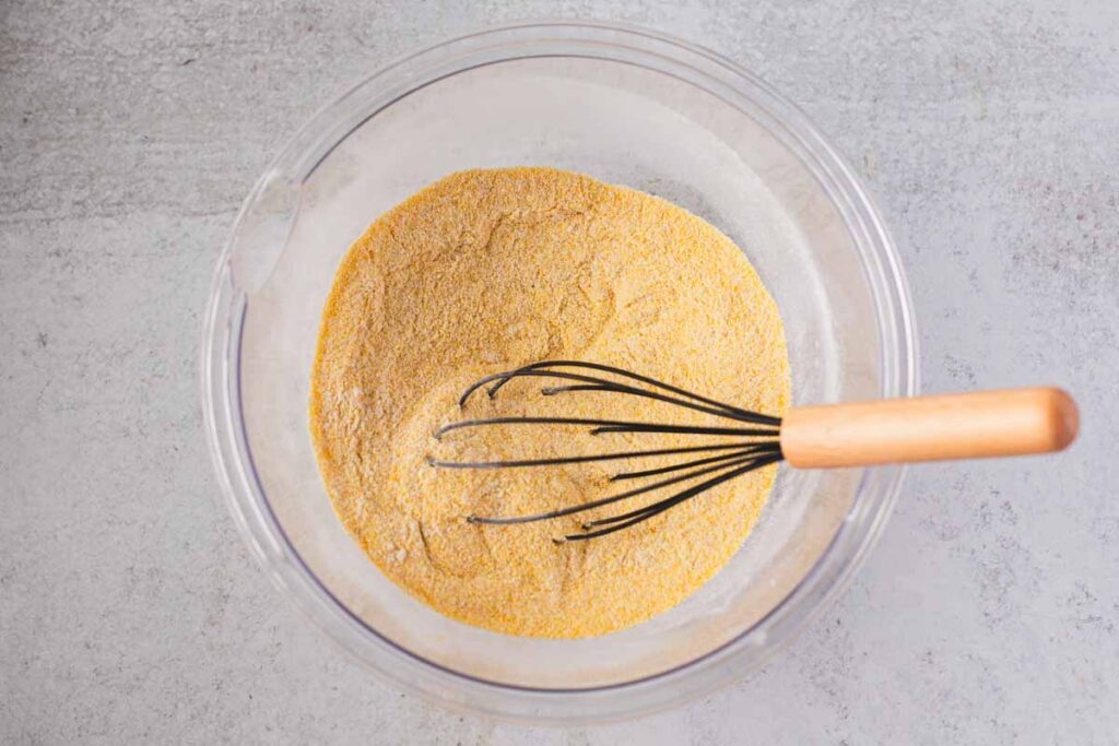 Cornmeal, flour, sugar, salt, baking powder, and baking soda whisked together in a glass bowl for skillet cornbread recipe.