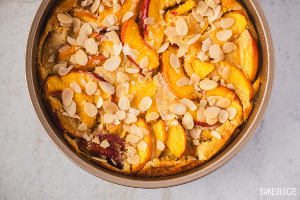 Baked peach clafoutis in a round pan, topped with sliced almonds and golden peach slices, fresh out of the oven.