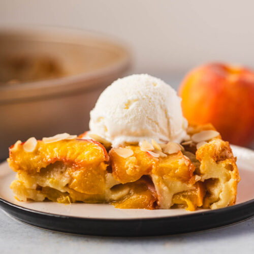 Slice of peach clafoutis topped with vanilla ice cream and sliced almonds on a plate, with a fresh peach and baking pan in the background.