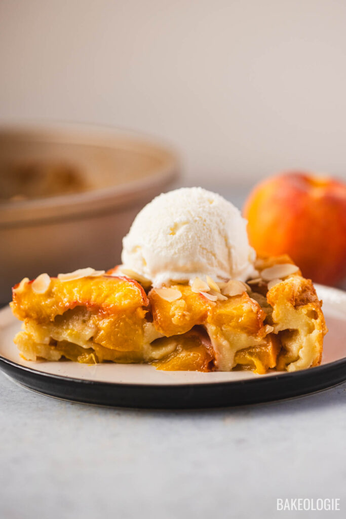 Slice of peach clafoutis topped with vanilla ice cream and sliced almonds on a plate, with a fresh peach and baking pan in the background.