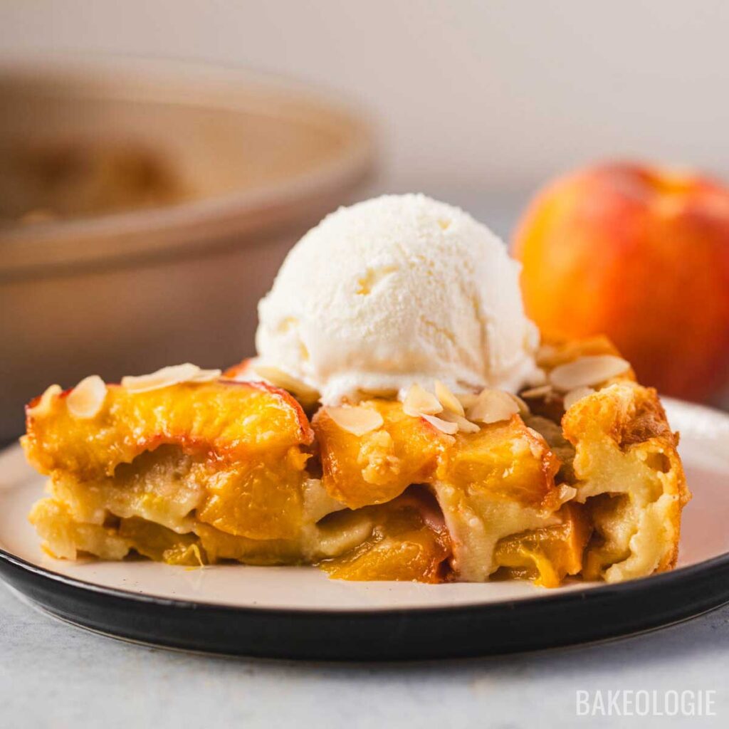 Slice of peach clafoutis topped with vanilla ice cream and sliced almonds on a plate, with a fresh peach and baking pan in the background.