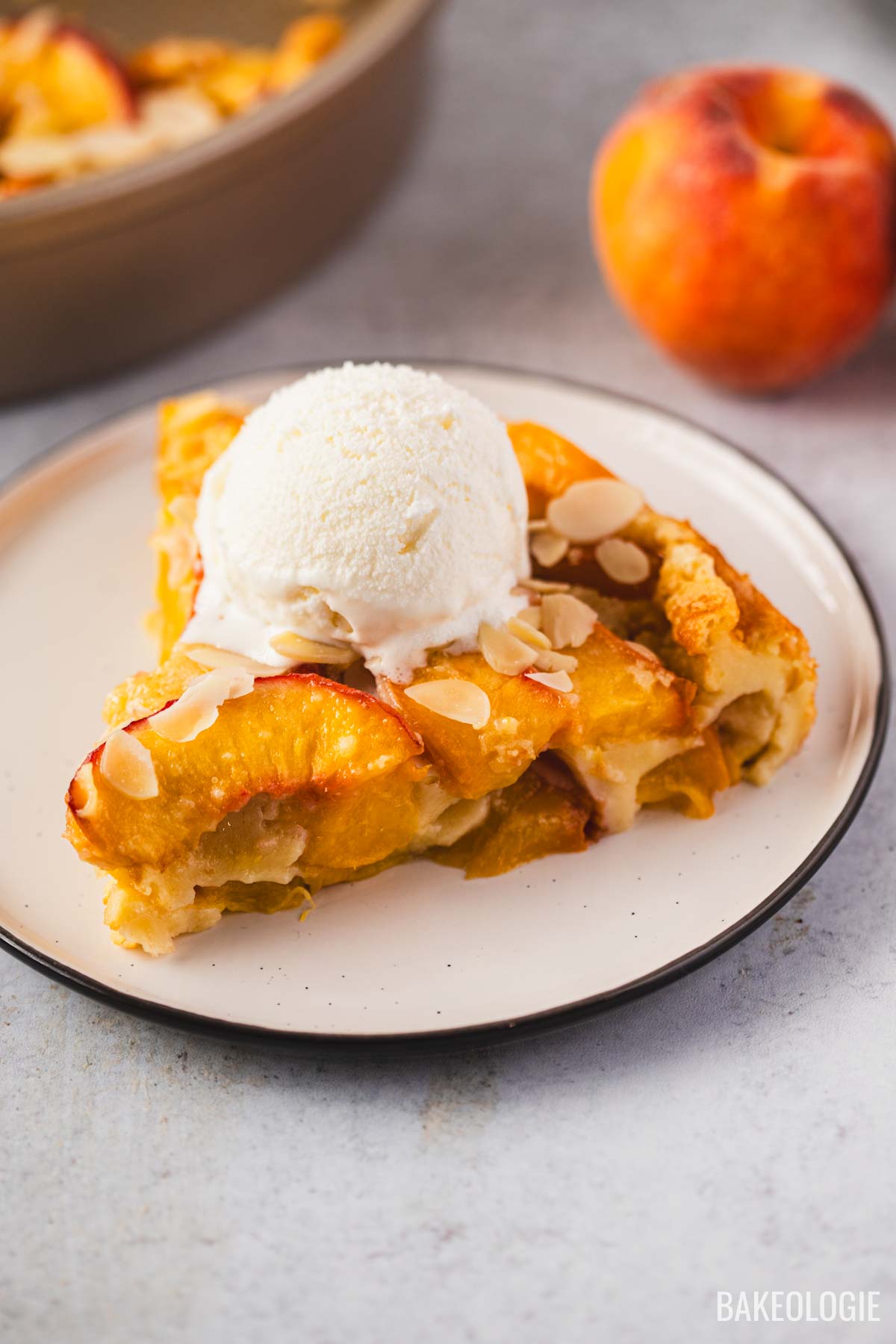 Slice of peach clafoutis on a plate, topped with a scoop of vanilla ice cream and sliced almonds, with a whole peach and baking pan in the background.