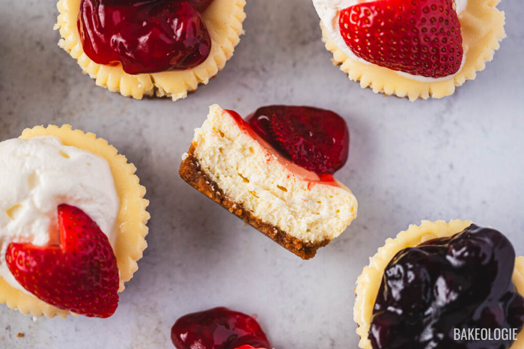 Assorted mini cheesecakes in muffin liners with graham cracker crust, topped with strawberries, cherries, and blueberries, one showing a creamy bite.