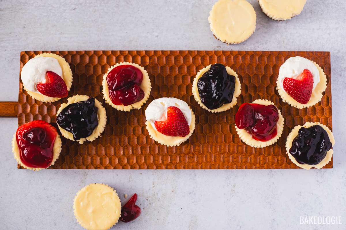 Mini cheesecakes baked in a muffin tin topped with strawberries, blueberry sauce, and cherry filling, displayed on a wooden tray.