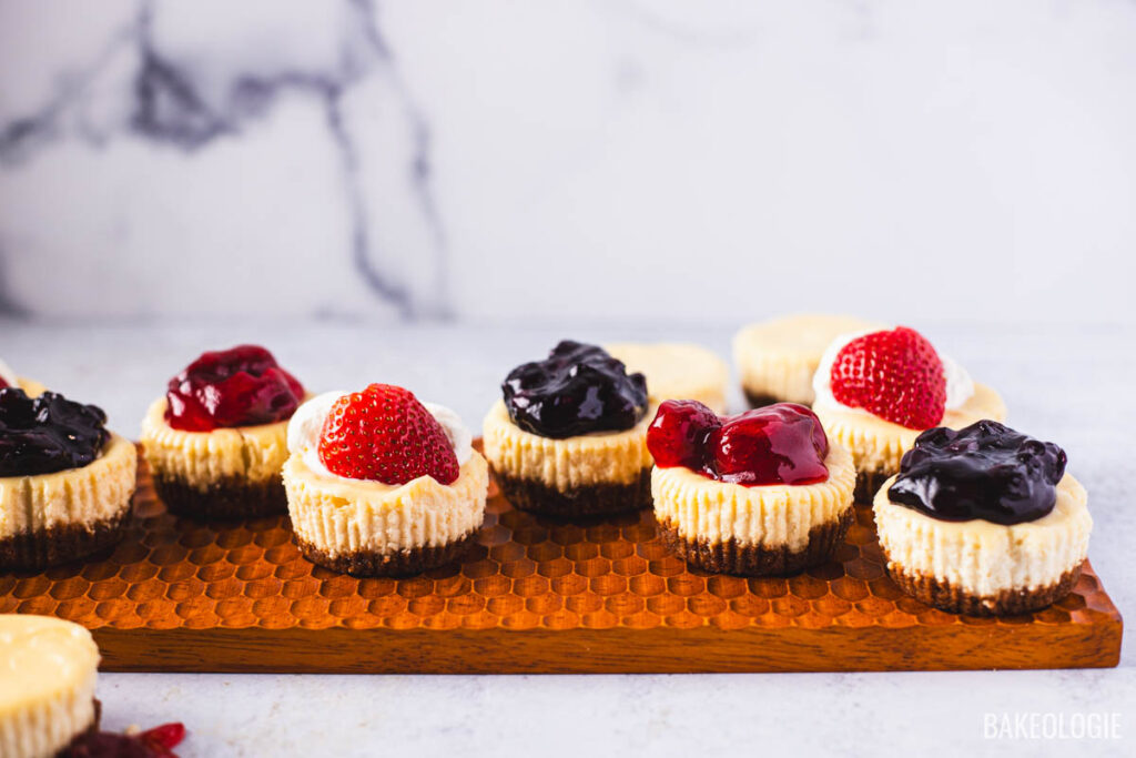 Assorted mini cheesecakes with graham cracker crust and different toppings like strawberry, blueberry, and cherry on a wooden tray.