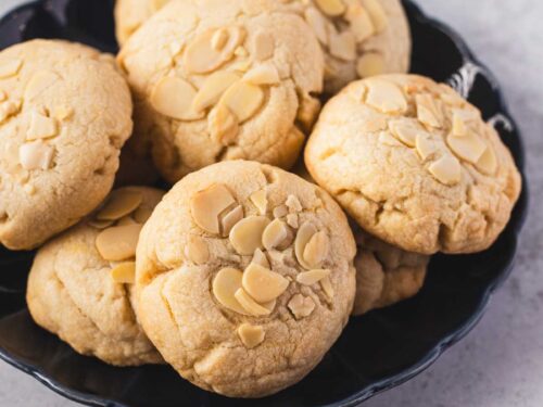 A plate of golden almond croissant cookies topped with sliced almonds, showing their soft, thick texture and slightly cracked tops.
