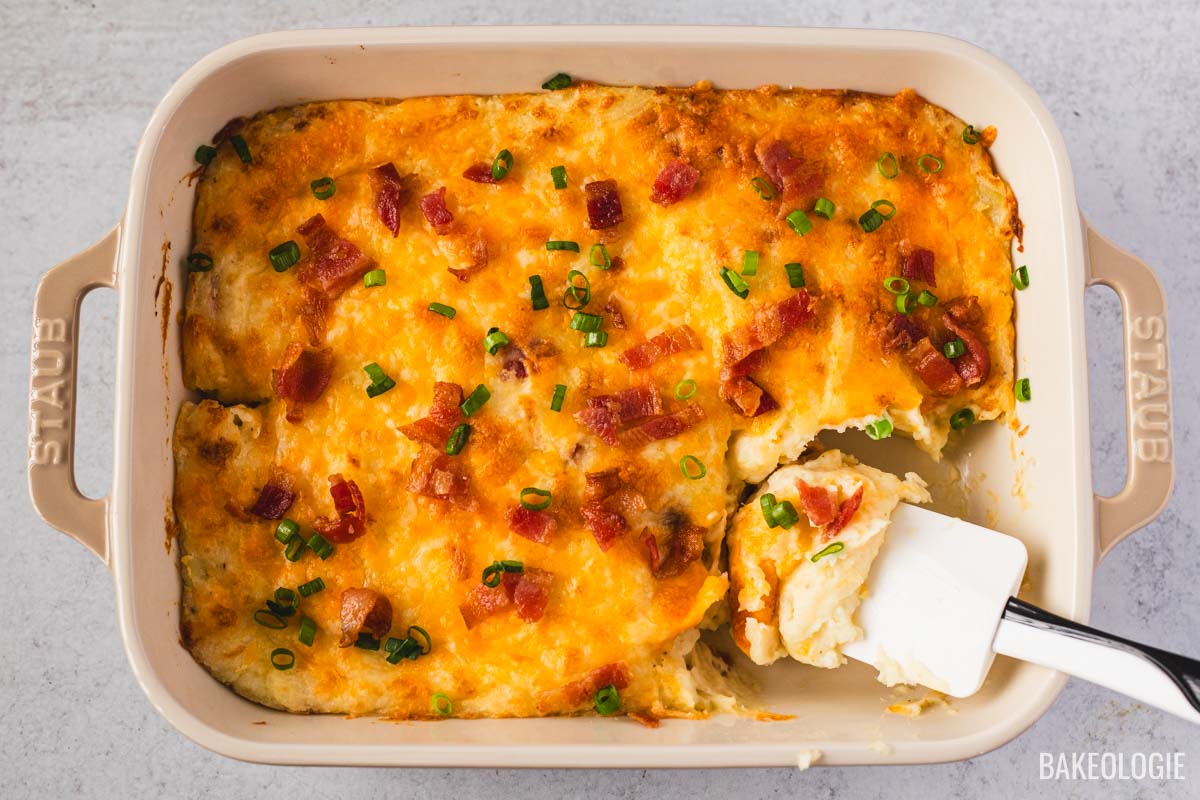 Overhead shot of a golden, cheesy loaded mashed potato casserole in a beige Staub baking dish. The top is sprinkled with crispy bacon and green onions, with a spatula lifting out a creamy, fluffy scoop.