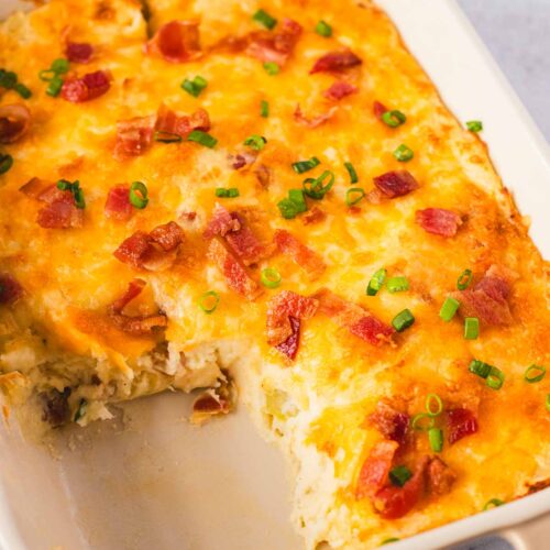 Angled view of a loaded mashed potato casserole with melted cheddar cheese, bacon bits, and chopped green onions on top. A portion has been scooped out, showing the creamy potato filling inside the beige Staub dish.