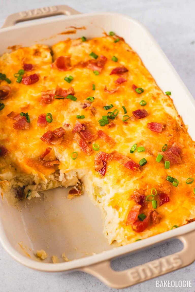 Angled view of a loaded mashed potato casserole with melted cheddar cheese, bacon bits, and chopped green onions on top. A portion has been scooped out, showing the creamy potato filling inside the beige Staub dish.