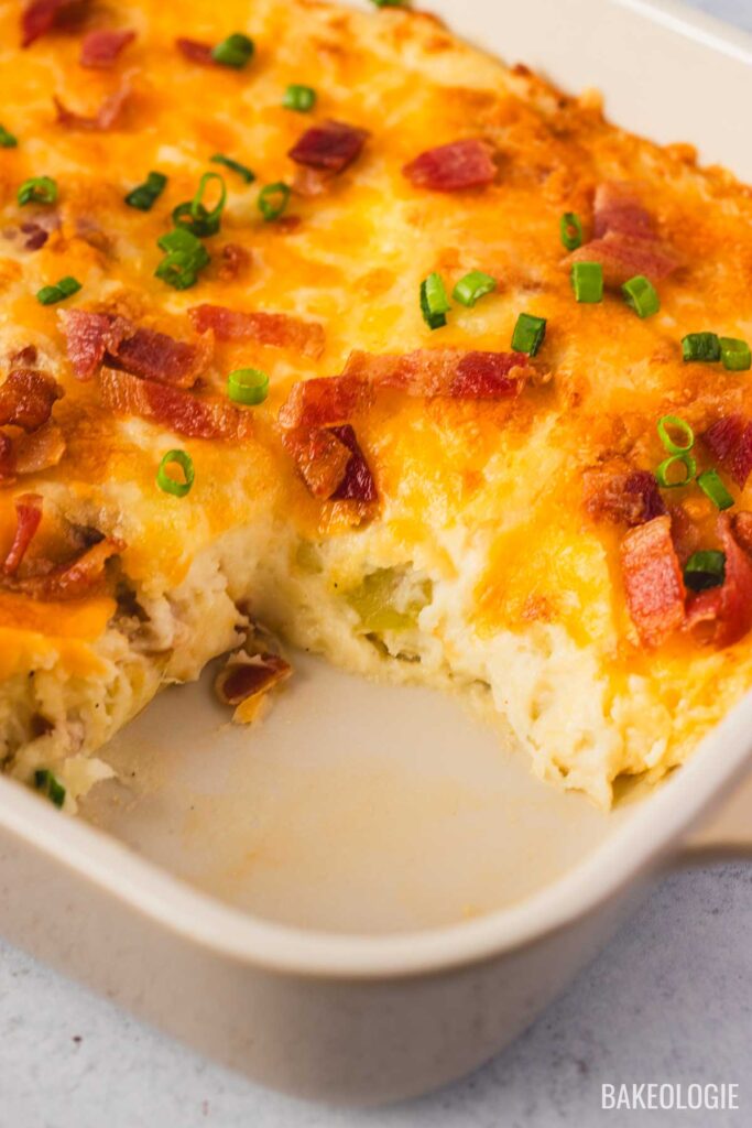 Close-up of the creamy mashed potato casserole inside a beige Staub baking dish. The golden melted cheese and bacon pieces are highlighted, with fresh green onions adding a pop of color on top.