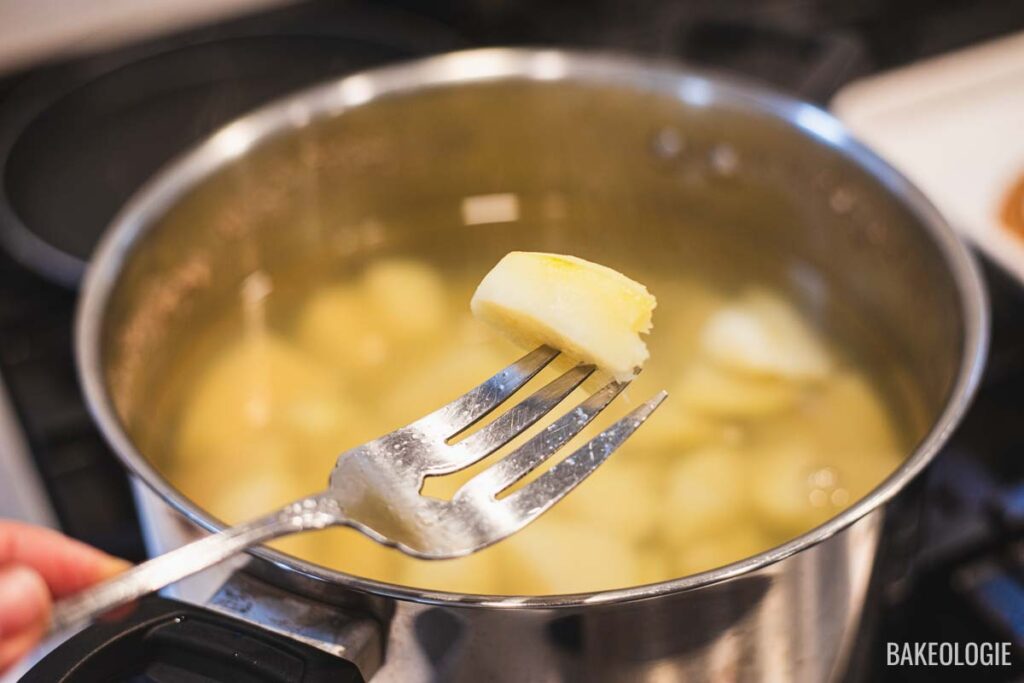 A fork testing a boiled potato cube for doneness over a pot of simmering potatoes on the stove.