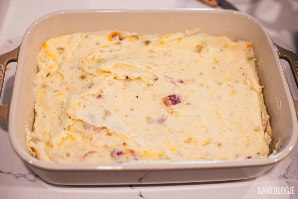 Mashed potato mixture with cheese and bacon spread evenly in a beige Staub baking dish before baking.