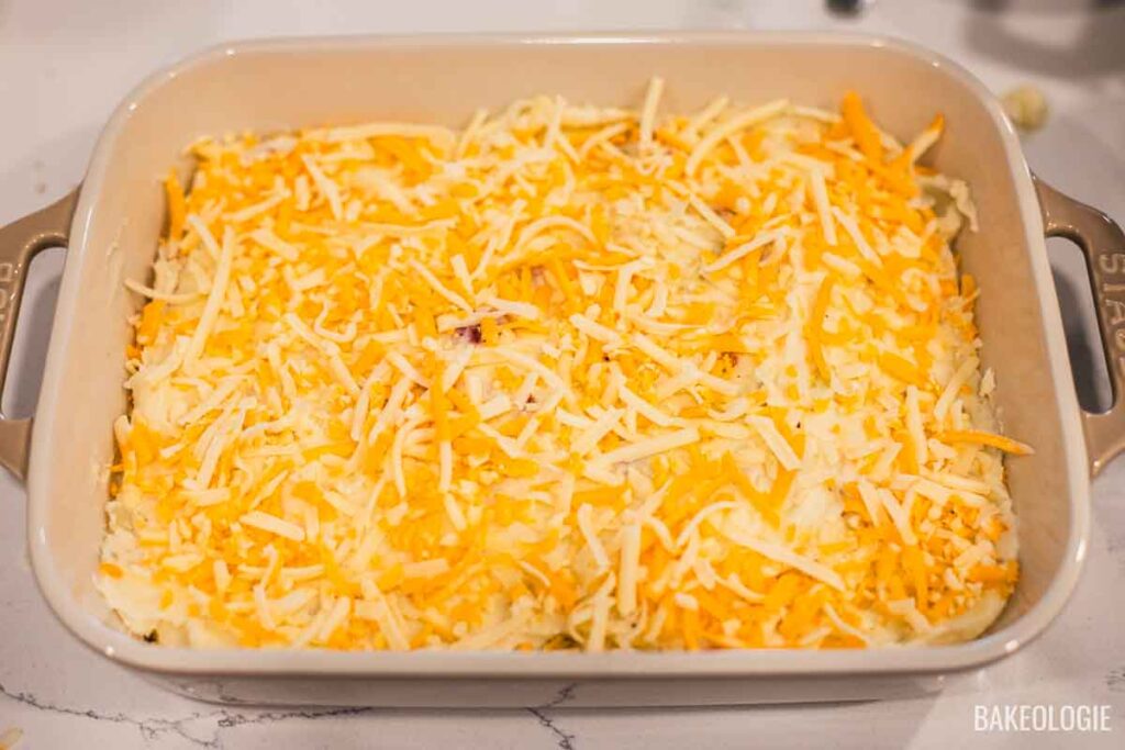 The mashed potato casserole topped generously with shredded cheddar and mozzarella cheese before baking.