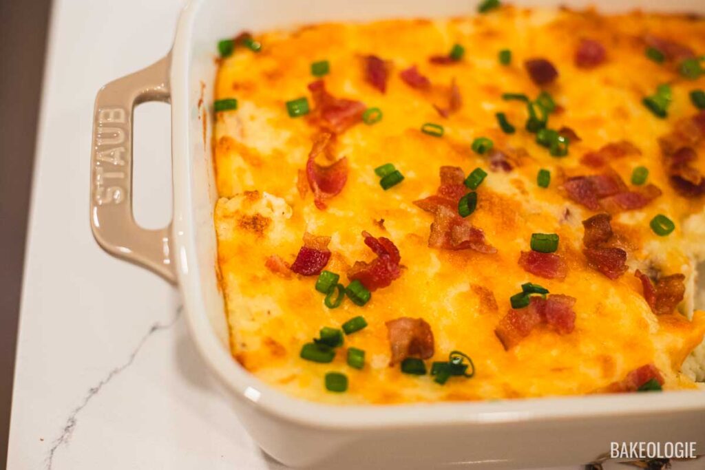 Freshly baked mashed potato casserole in a beige Staub dish, topped with melted cheese, crispy bacon, and green onions.
