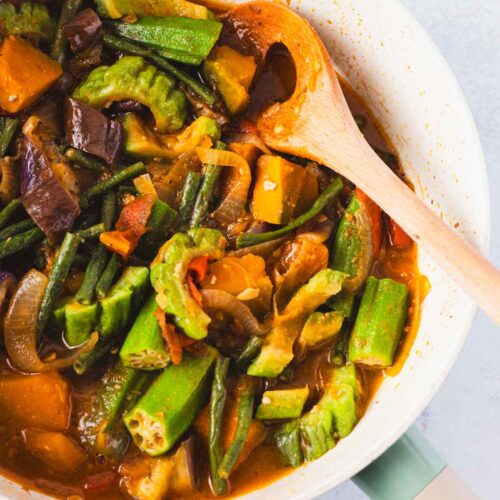 Overhead view of a Filipino Pinakbet recipe in a pan with a wooden spoon, showing tender okra, long beans, squash, eggplant, and bitter melon in a savory shrimp paste sauce.