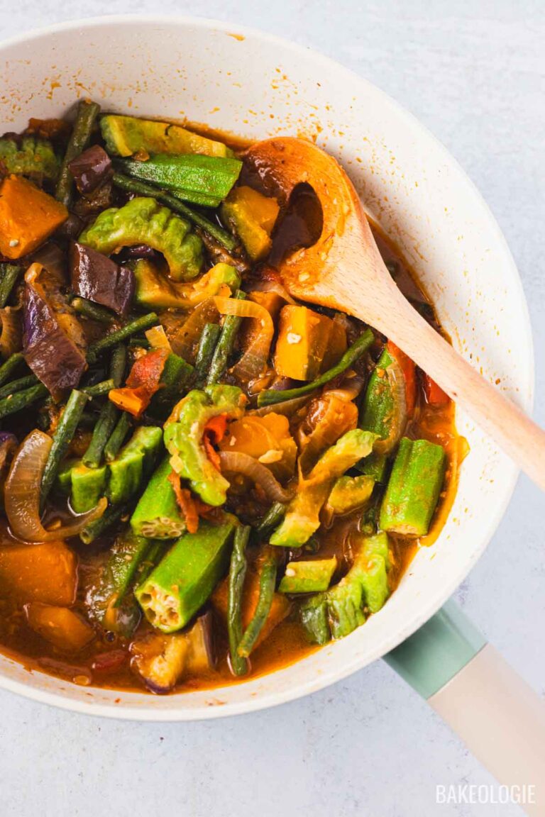 Overhead view of a Filipino Pinakbet recipe in a pan with a wooden spoon, showing tender okra, long beans, squash, eggplant, and bitter melon in a savory shrimp paste sauce.