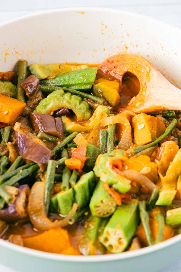 Overhead view of a Filipino Pinakbet recipe in a pan with a wooden spoon, showing tender okra, long beans, squash, eggplant, and bitter melon in a savory shrimp paste sauce.