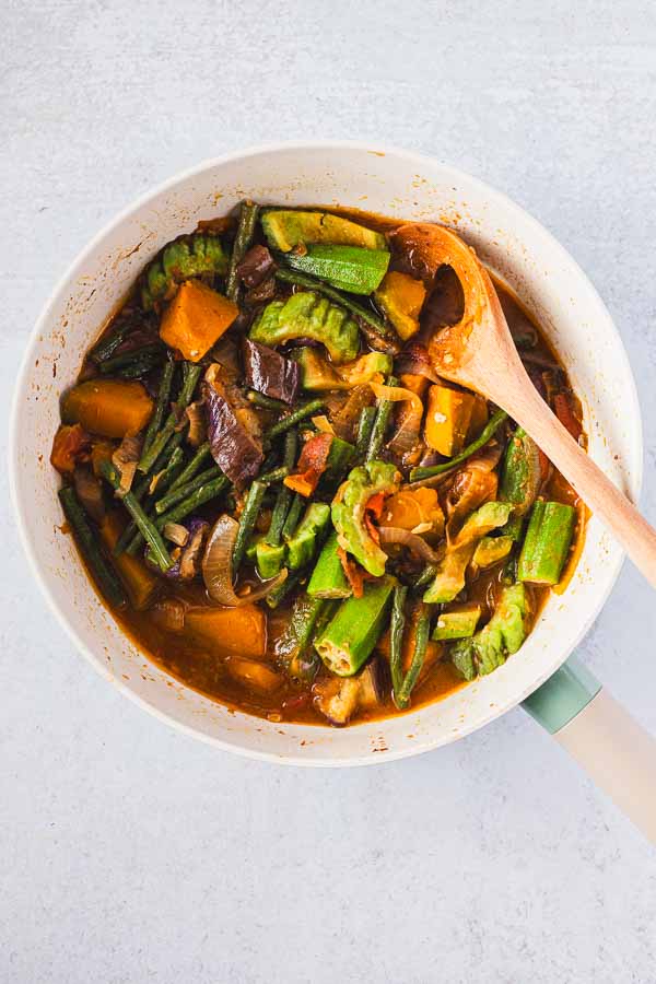 Overhead view of a Filipino Pinakbet recipe in a pan with a wooden spoon, showing tender okra, long beans, squash, eggplant, and bitter melon in a savory shrimp paste sauce.