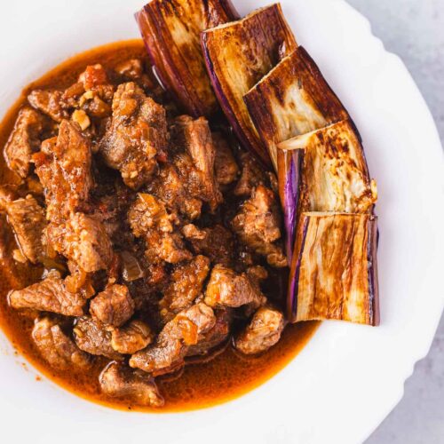 Overhead shot of binagoongang baboy in a white bowl, with rich shrimp paste sauce and fried eggplant slices.