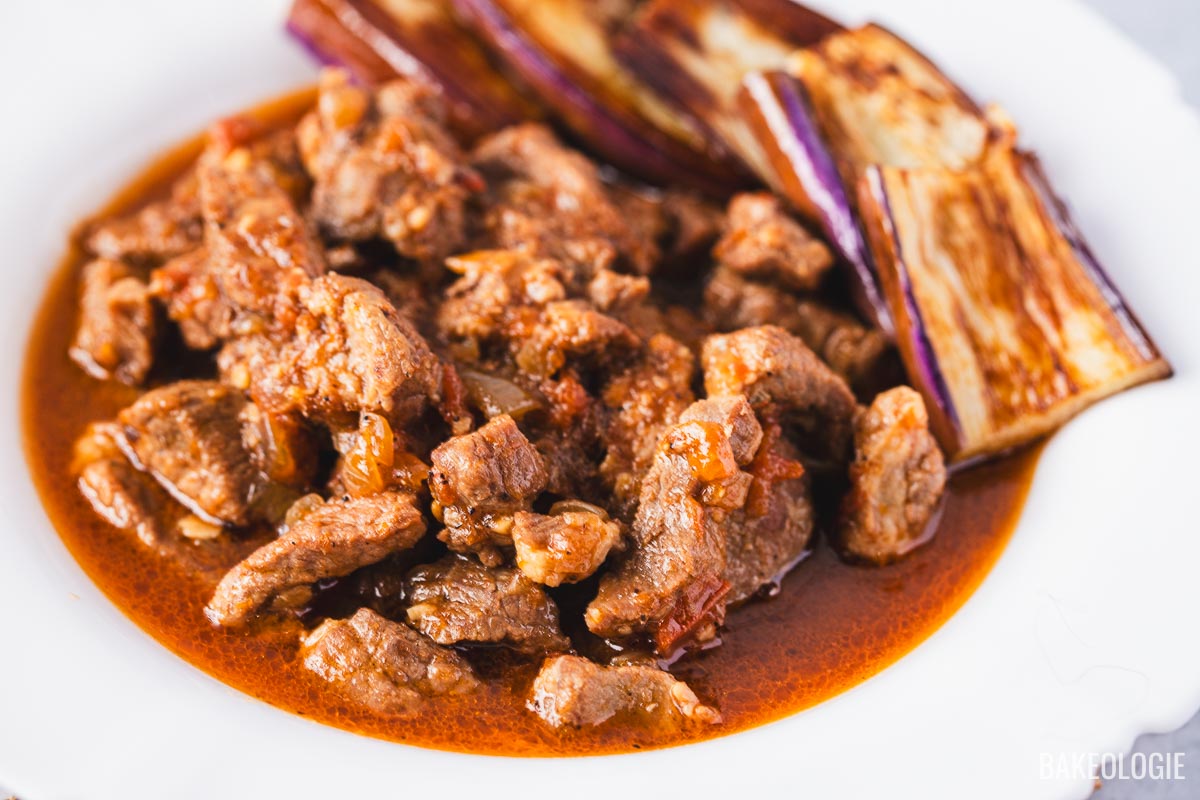 Close-up of Filipino binagoongang baboy, tender pork cooked in shrimp paste sauce, served with fried eggplant on the side.