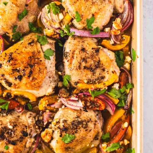 Close-up of sheet pan Greek chicken thighs surrounded by roasted vegetables, lemon slices, and feta for an easy one-pan Mediterranean dinner.