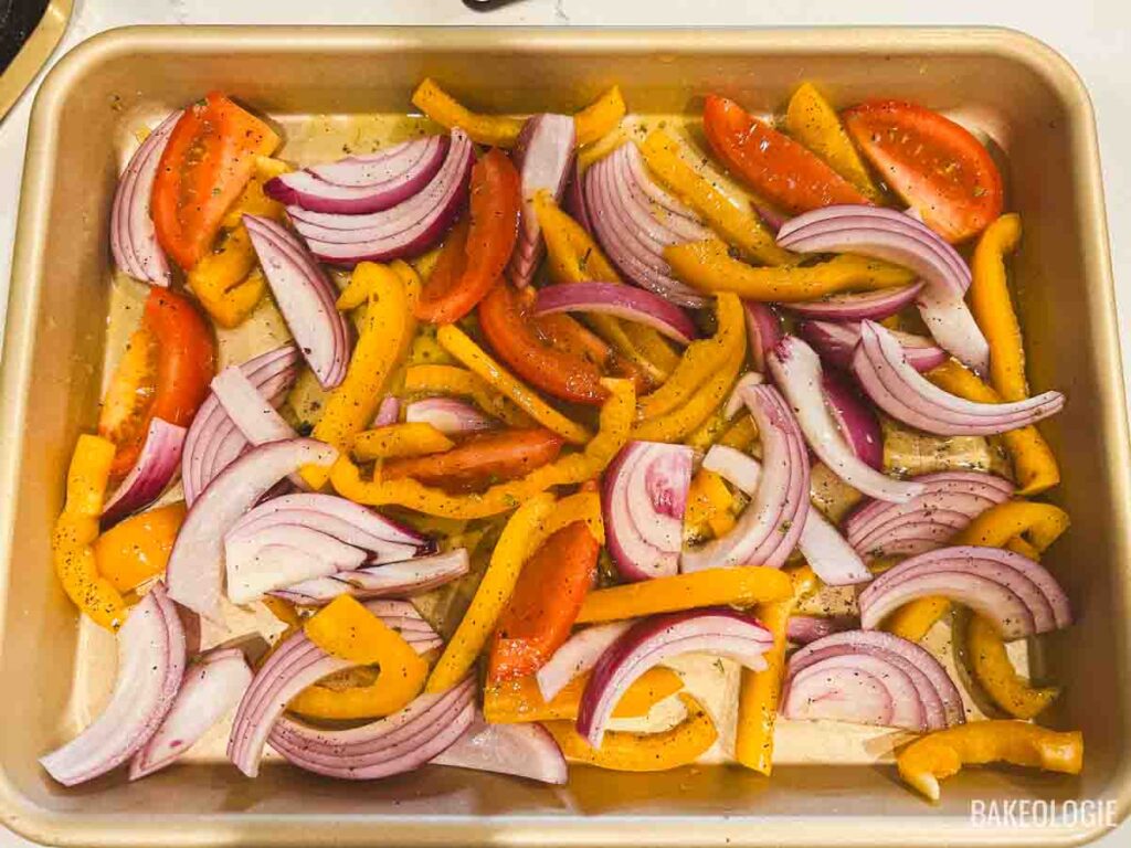 Bell peppers, red onions, and tomato wedges arranged on a sheet pan and coated with Greek lemon-oregano marinade.