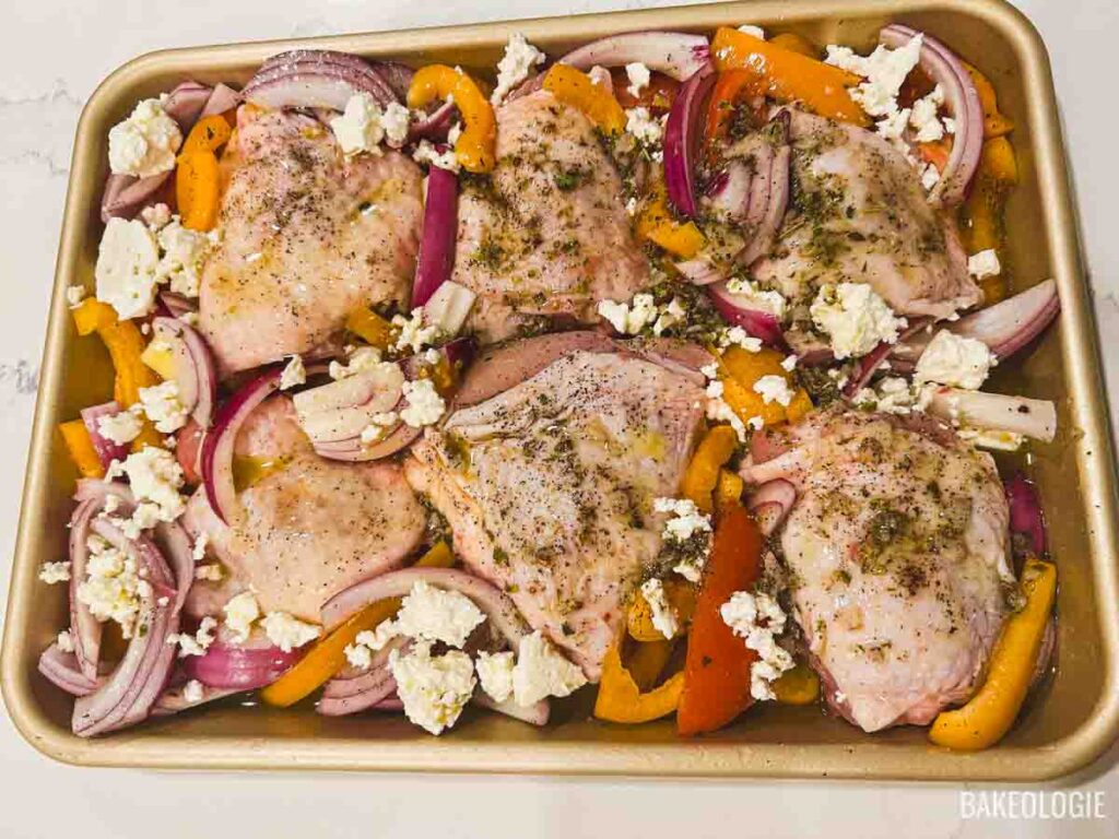 Raw chicken thighs placed between seasoned vegetables on a sheet pan and drizzled with Greek marinade, ready for the oven.