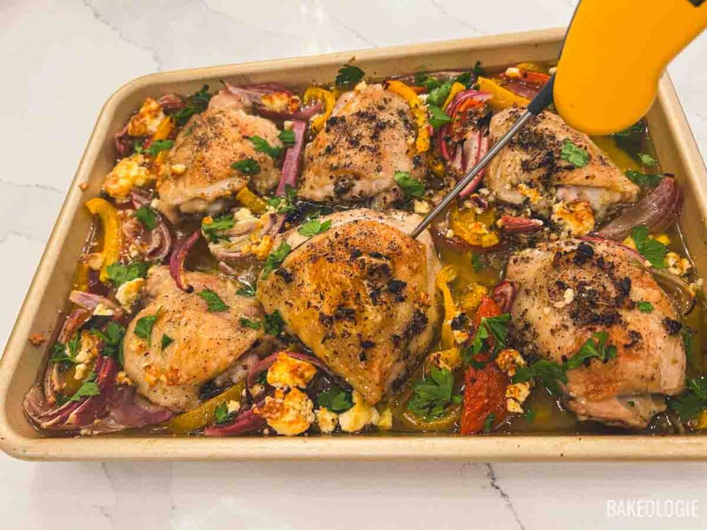 Sheet pan Greek chicken just out of the oven, golden and juicy, being checked with a thermometer to ensure perfect doneness.