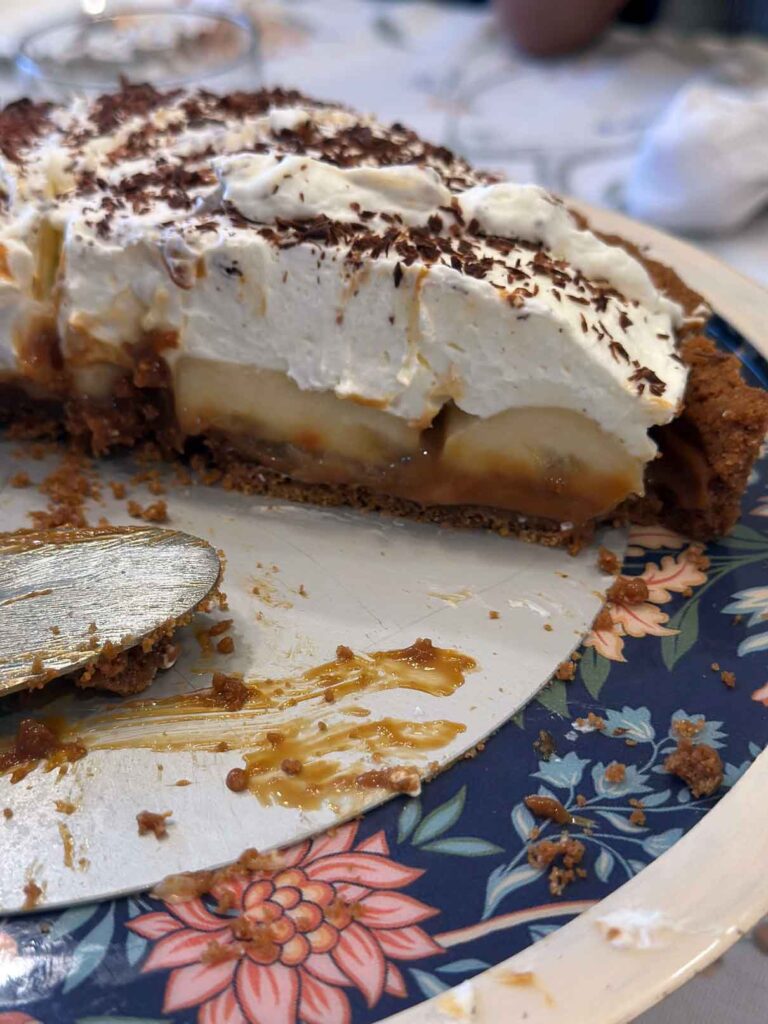 Slice of homemade banoffee pie showing layers of graham cracker crust, caramel, fresh bananas, whipped cream, and chocolate shavings on top.