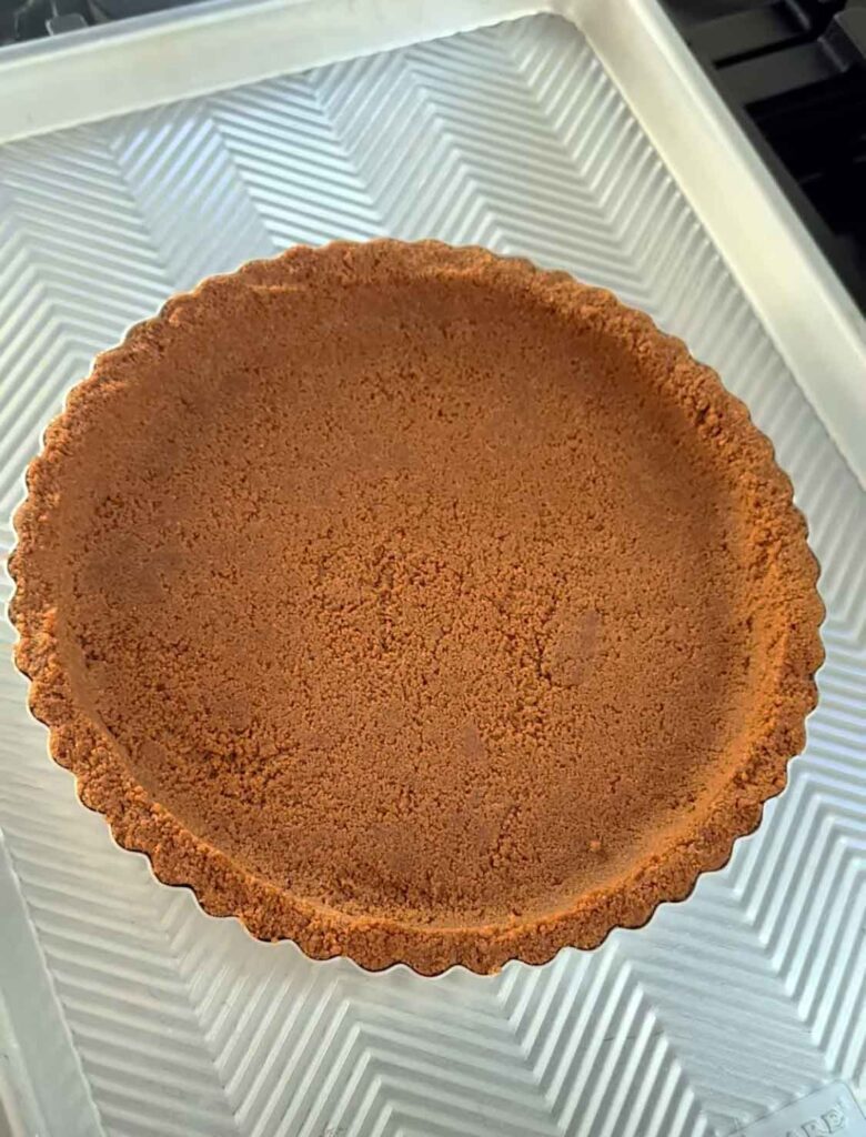Baked graham cracker crust in a fluted 9-inch tart pan, golden brown with neat, compact sides and even crumb base, cooling on a baking sheet.