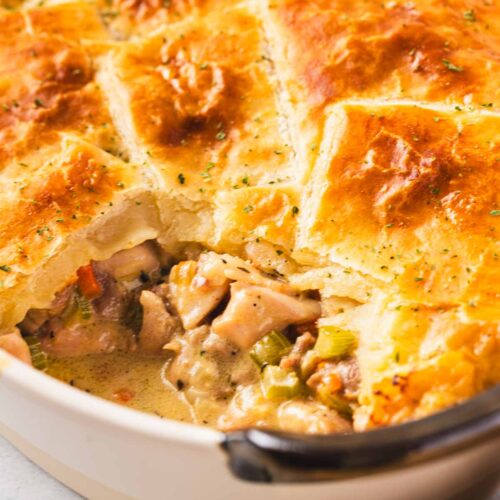 Close-up of a creamy chicken pot pie topped with golden puff pastry squares in a white baking dish, showing the tender chicken and vegetables in a rich sauce.